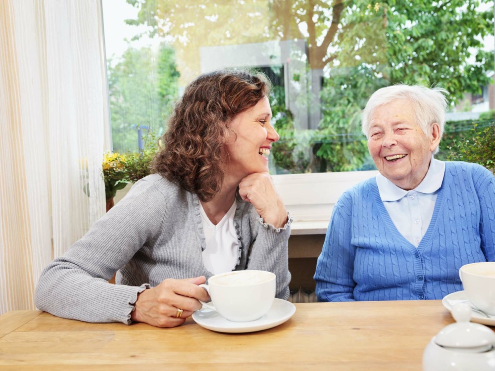 caregiver drinking coffee and talking with senior woman, having some fun