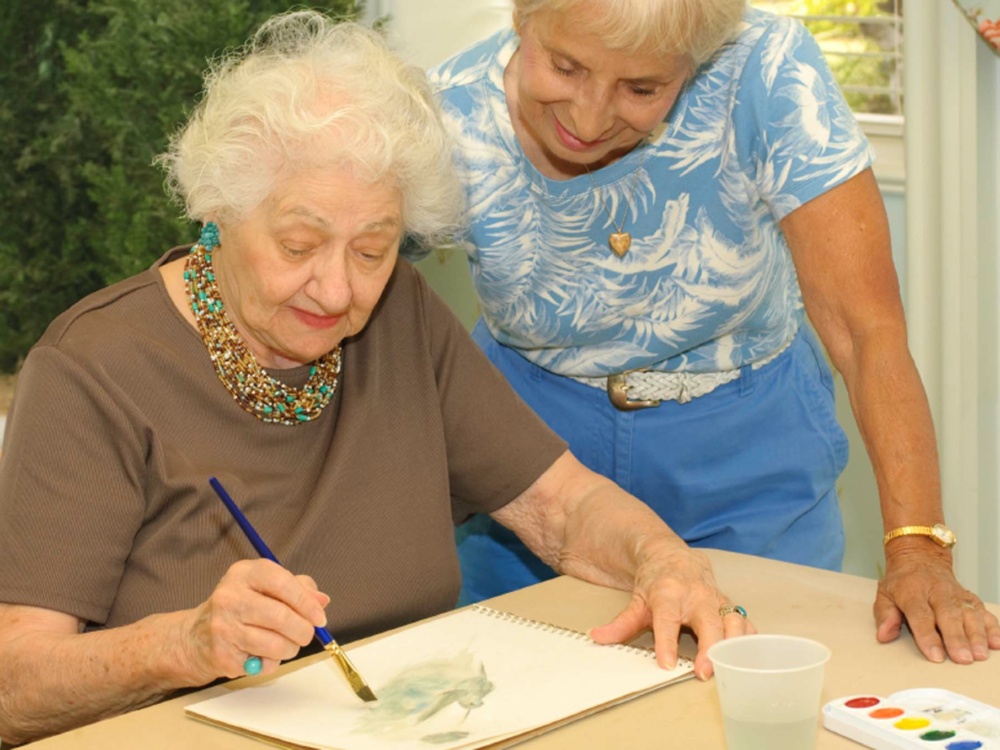 The older friend, 92, demonstrates her painting technique for her younger (84 year old) friend.